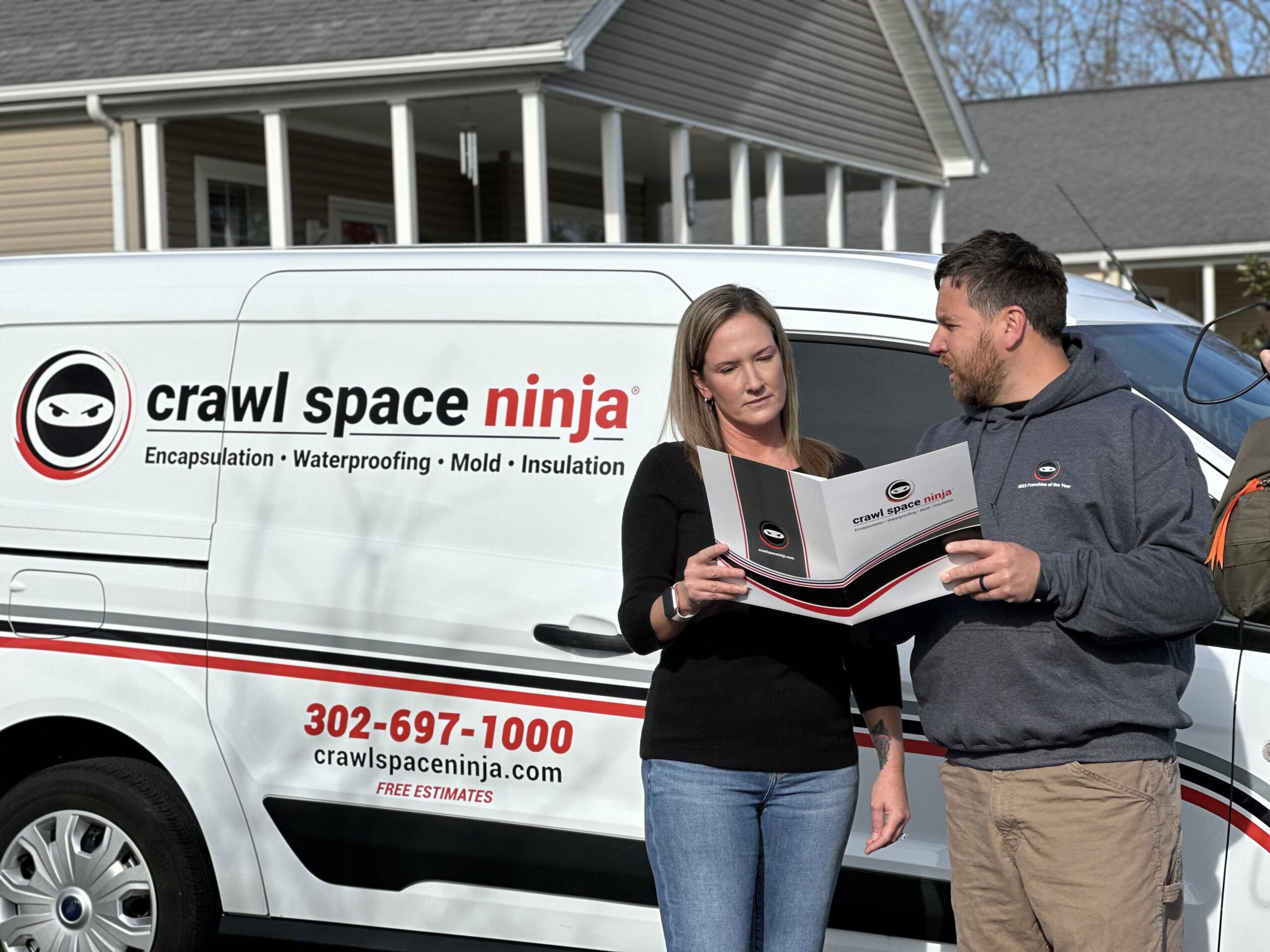 Two people stand beside a white van with a Crawl Space Ninja logo and service list, reading a brochure.