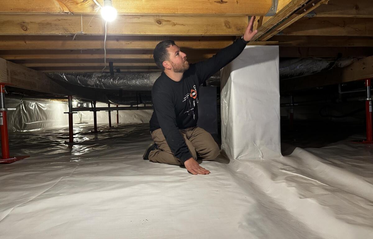 Man kneeling on a plastic-covered basement floor, reaching up to a wooden floor joist under a bare hanging light bulb