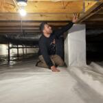 Man kneeling on a plastic-covered basement floor, reaching up to a wooden floor joist under a bare hanging light bulb