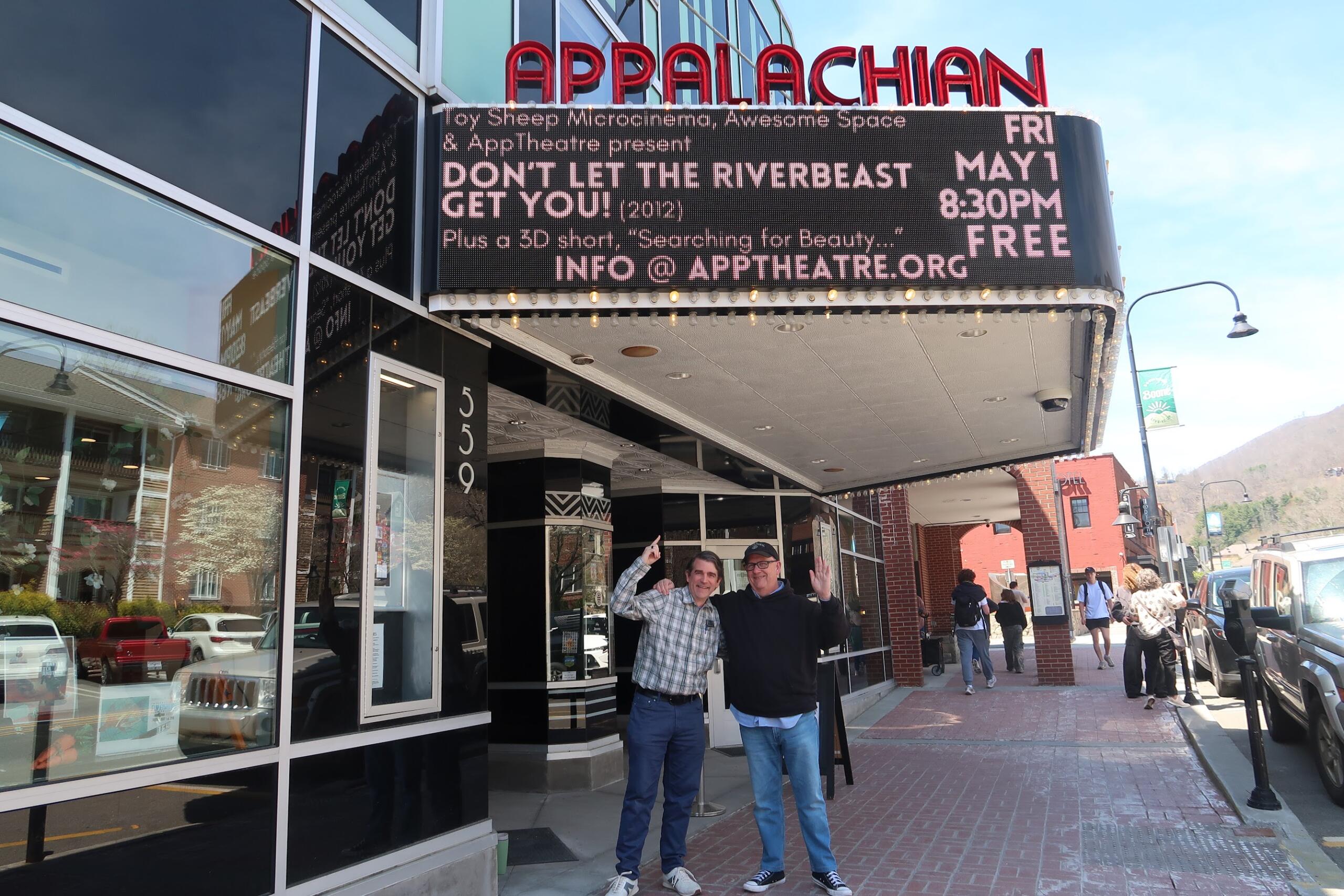 Two men stand on a brick sidewalk in front of a theater marquee, smiling and waving at the camera.