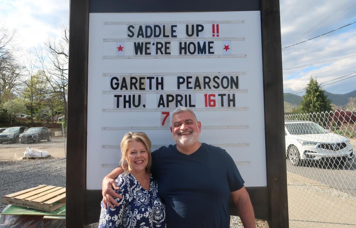 Couple poses in front of a white signboard announcing a horse-riding event, smiling with arms around each other.