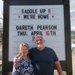 Couple poses in front of a white signboard announcing a horse-riding event, smiling with arms around each other.