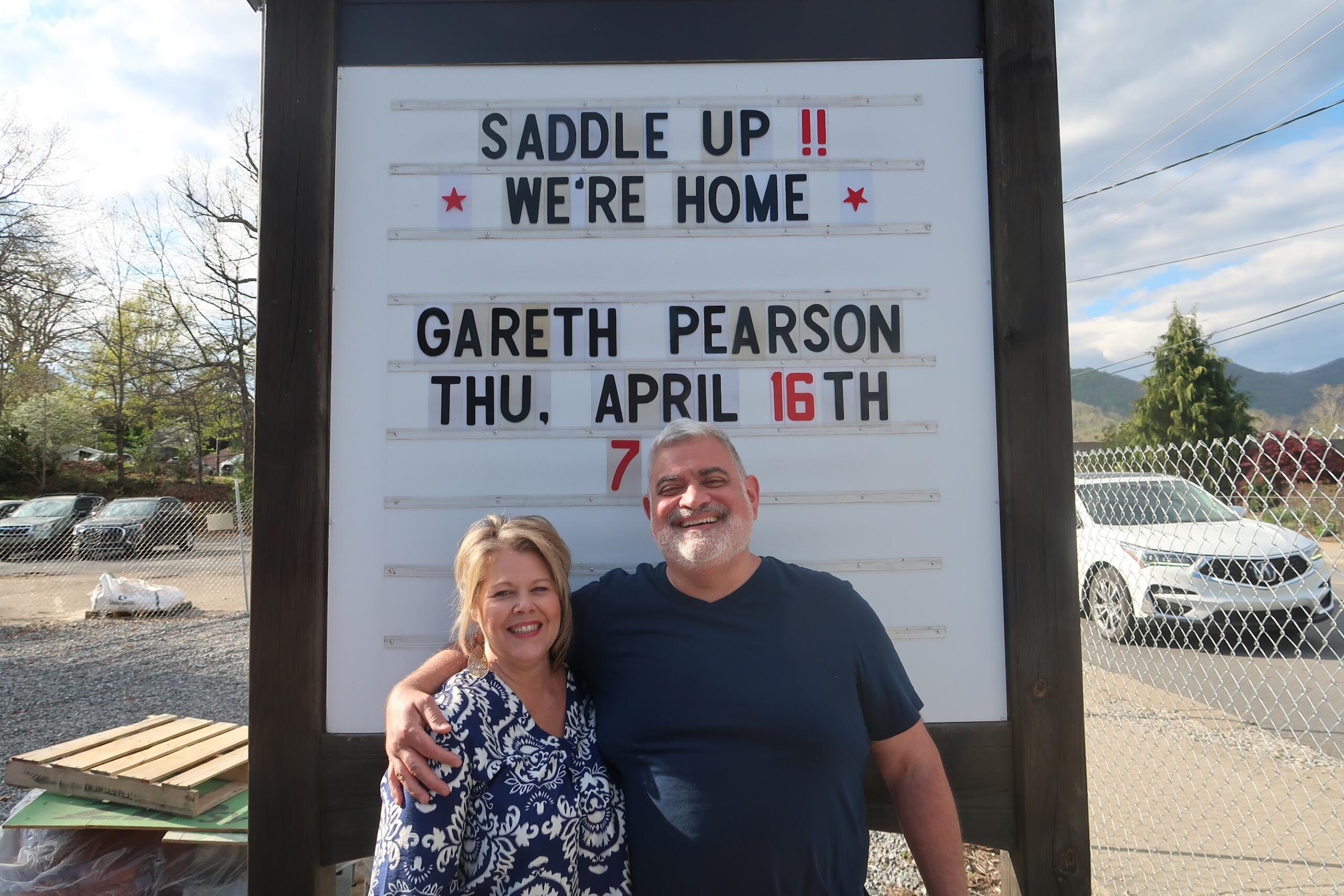 Couple poses in front of a white signboard announcing a horse-riding event, smiling with arms around each other.