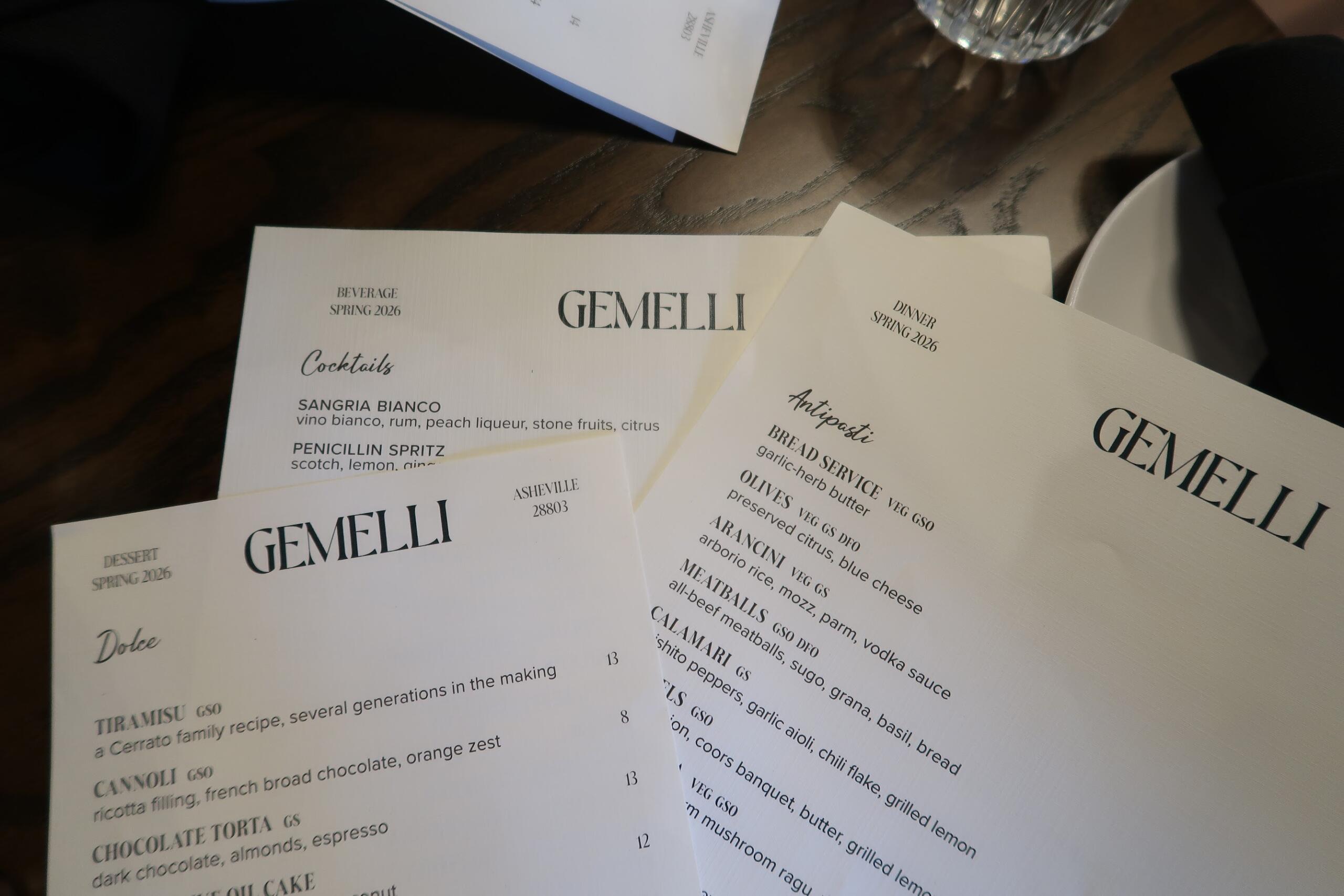 Group of white menu cards spread on a dark wooden table, reading GEMELLI and Spring 2026 cocktails and dinner sections (restaurant menus).