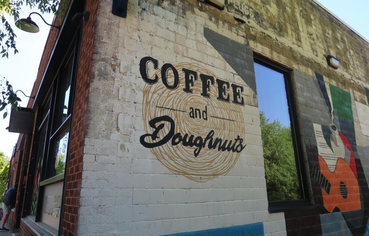 Brick storefront with mural reading 'Coffee and Donuts' over a circular swirl; large window to the right reflecting trees.