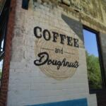Brick storefront with mural reading 'Coffee and Donuts' over a circular swirl; large window to the right reflecting trees.