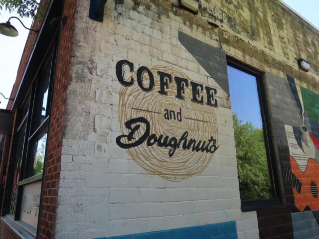 Brick storefront with mural reading 'Coffee and Donuts' over a circular swirl; large window to the right reflecting trees.
