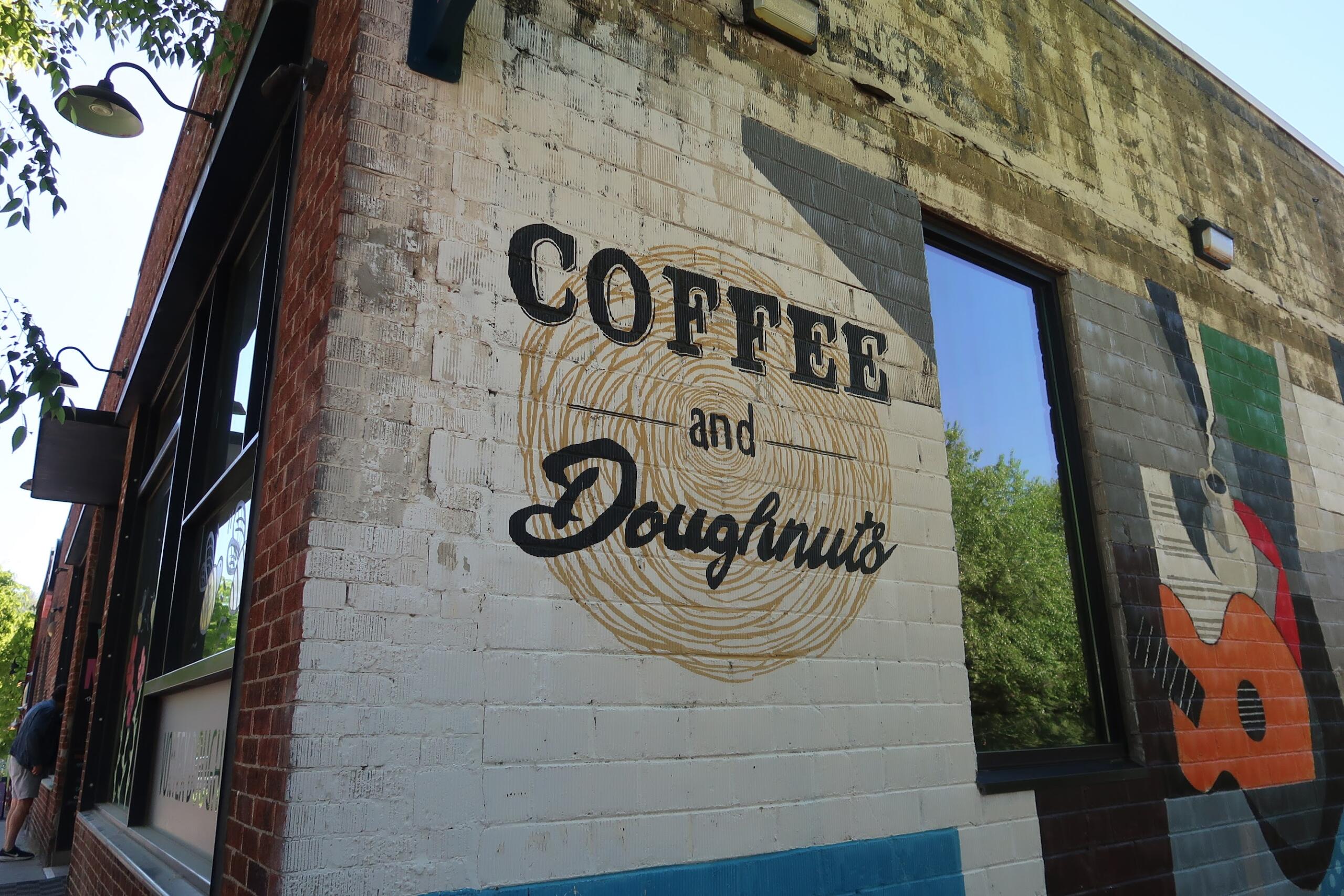 Brick storefront with mural reading 'Coffee and Donuts' over a circular swirl; large window to the right reflecting trees.