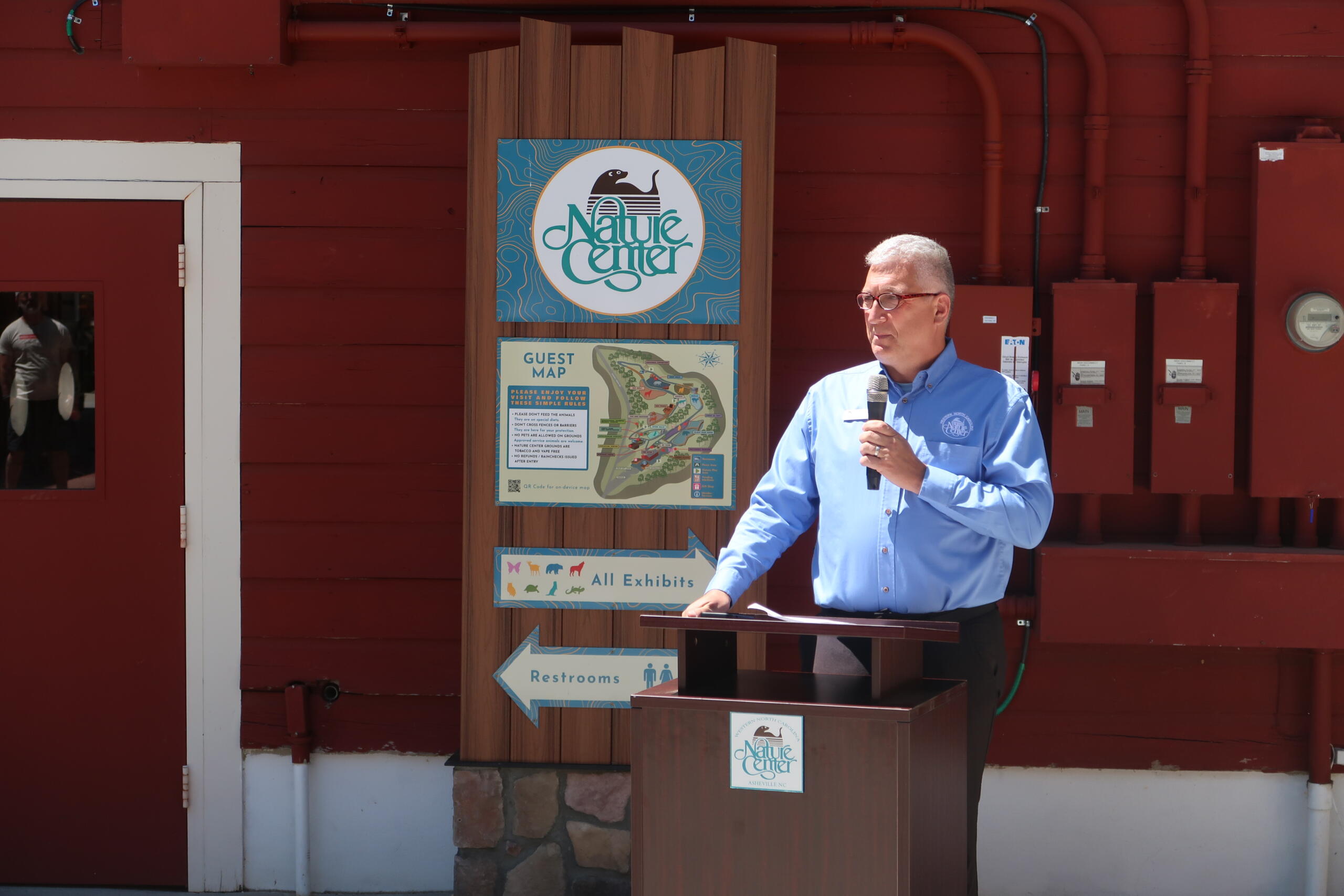 A man in a blue shirt speaks into a handheld microphone at a wooden podium outside a nature center signboard.