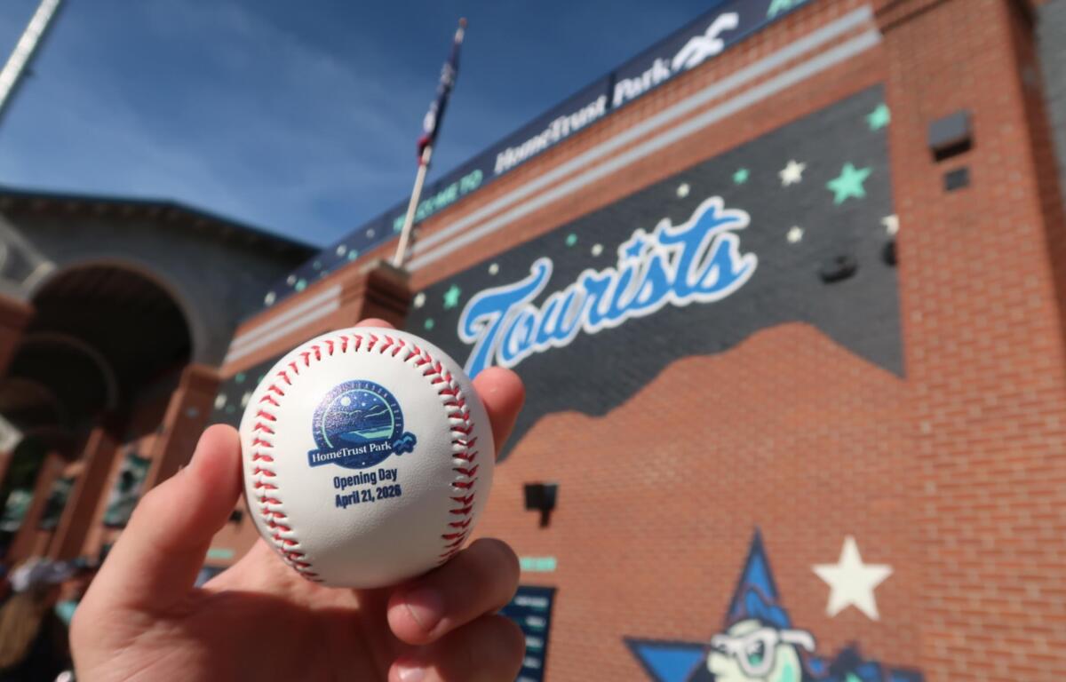 Close-up of a baseball with the HomeTrust Park Opening Day logo and date, held in front of a brick stadium wall.