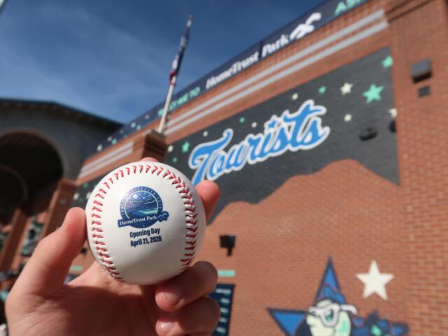 Close-up of a baseball with the HomeTrust Park Opening Day logo and date, held in front of a brick stadium wall.