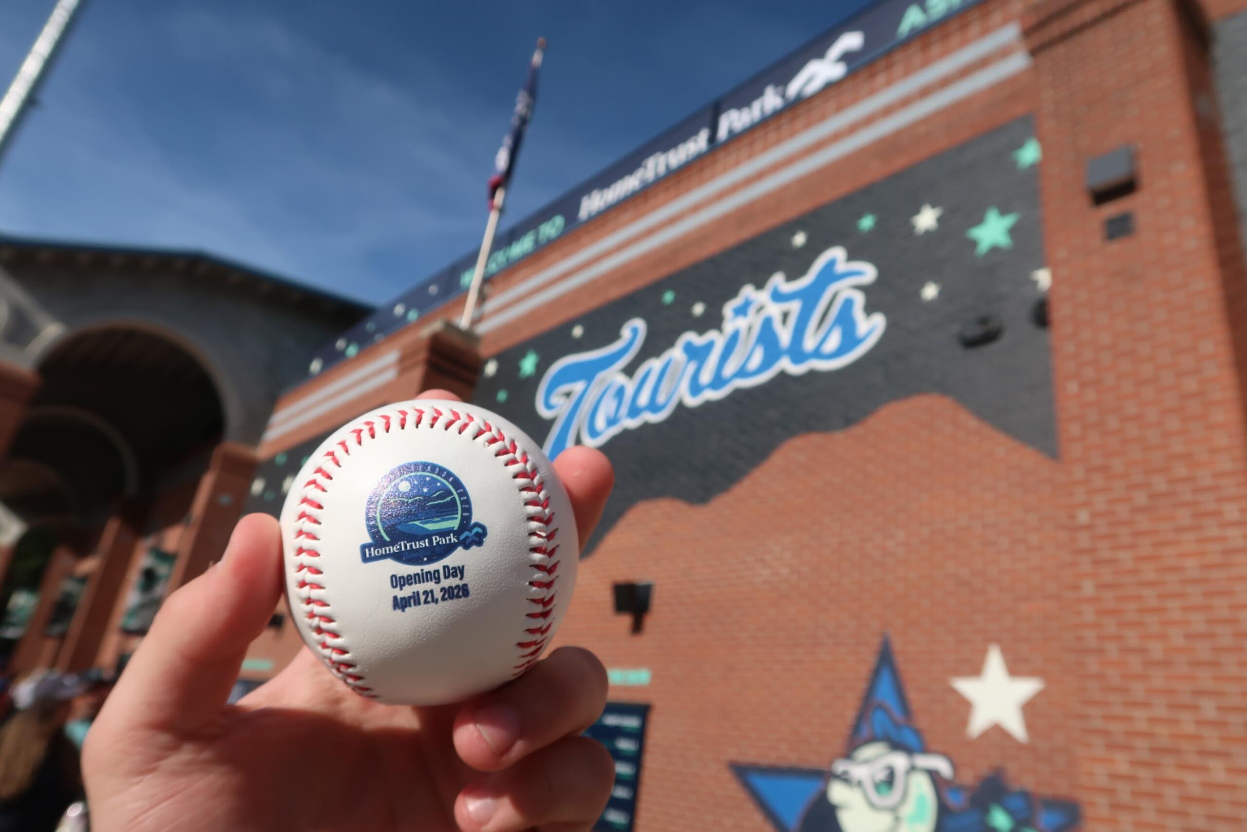 Close-up of a baseball with the HomeTrust Park Opening Day logo and date, held in front of a brick stadium wall.