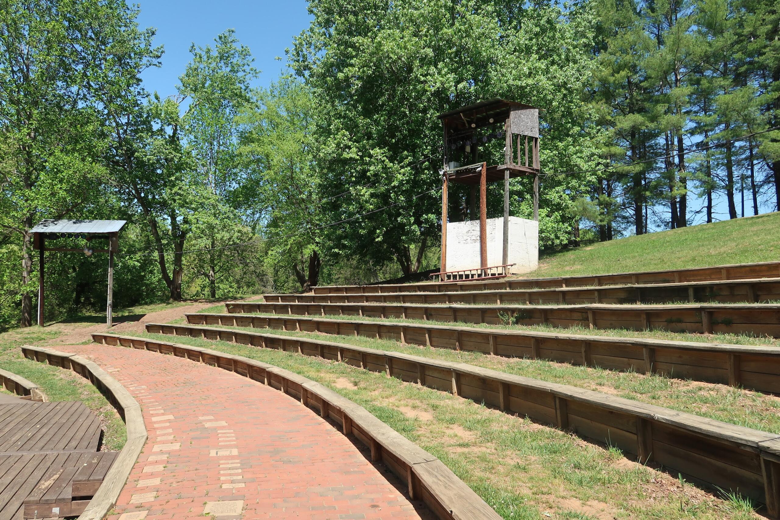 Outdoor park amphitheater with brick steps and tiered wooden seating, surrounded by trees and a tall wooden tower on the right.