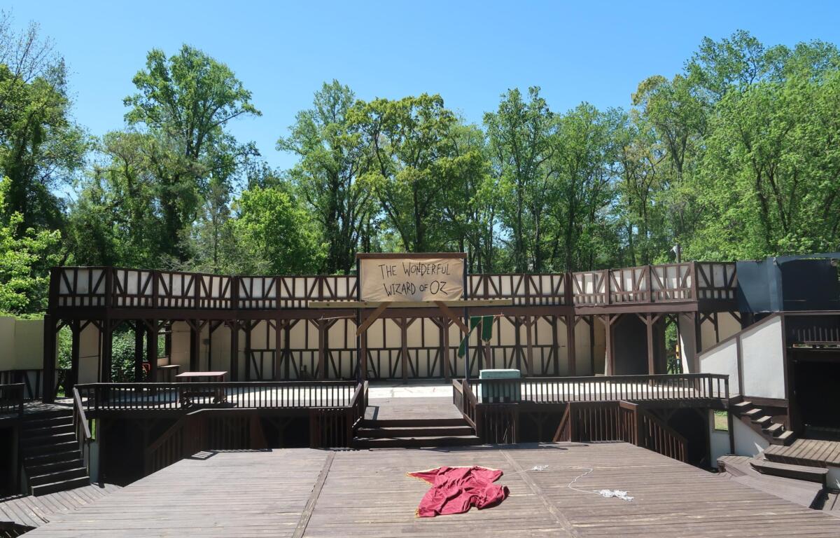 Outdoor wooden stage with a banner reading 'The Wonderful Wizard of Oz' against a backdrop of green trees and a clear blue sky.