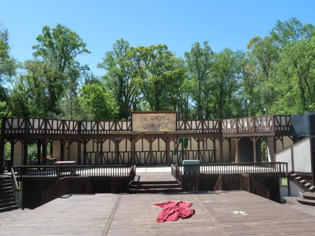Outdoor wooden stage with a banner reading 'The Wonderful Wizard of Oz' against a backdrop of green trees and a clear blue sky.