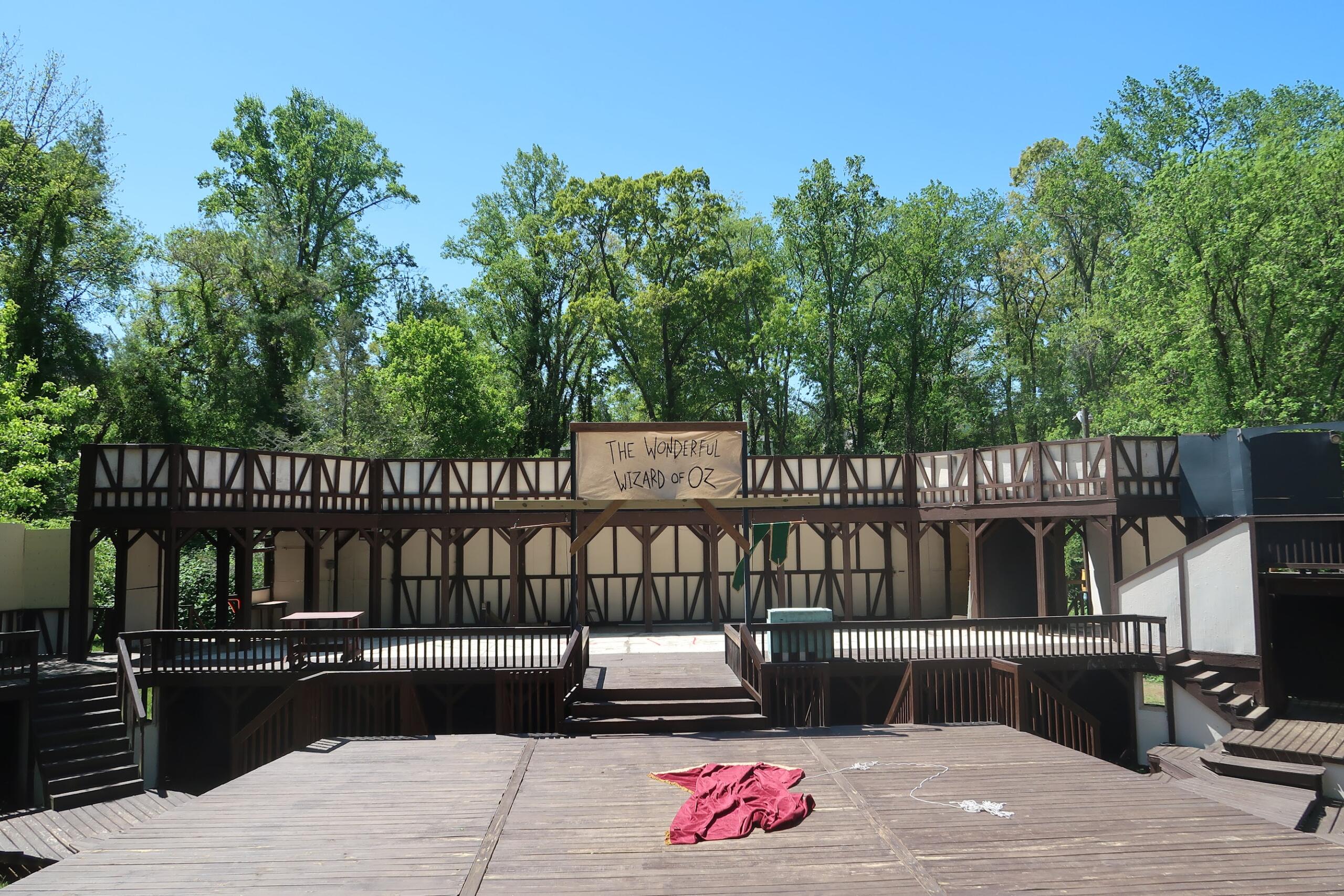 Outdoor wooden stage with a banner reading 'The Wonderful Wizard of Oz' against a backdrop of green trees and a clear blue sky.