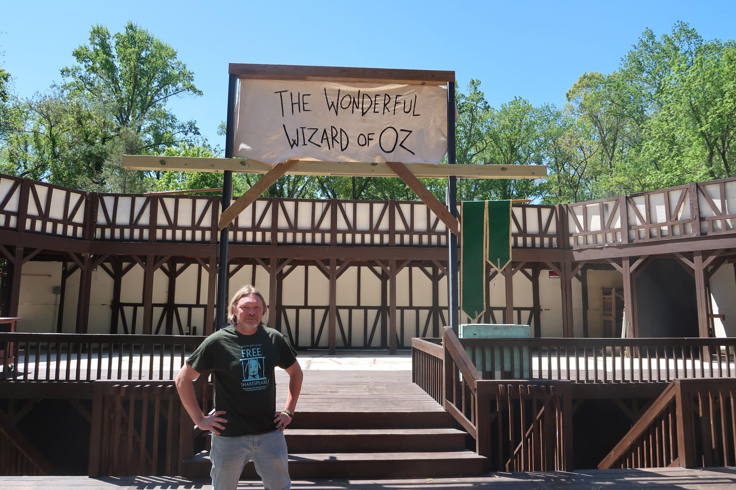Man posing in front of an outdoor wooden stage with a banner that says The Wonderful Wizard of Oz.
