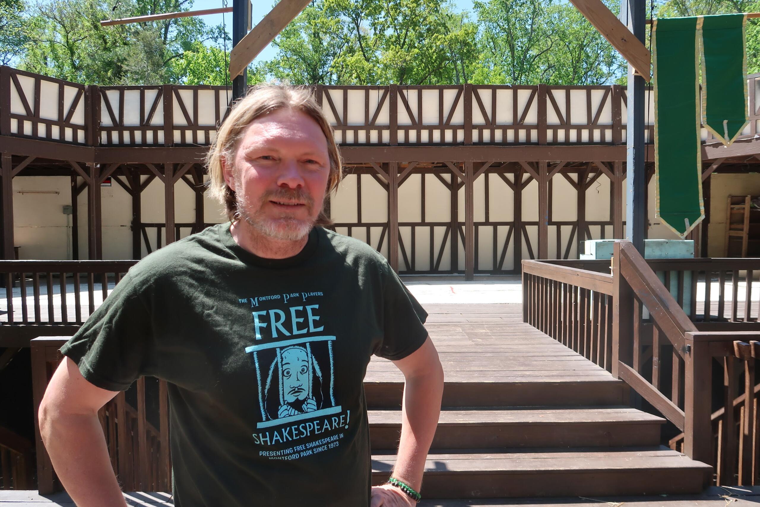 Man with light brown hair and a beard, wearing a dark green Shakespeare-themed T-shirt, standing on a wooden deck with brown railings outdoors.