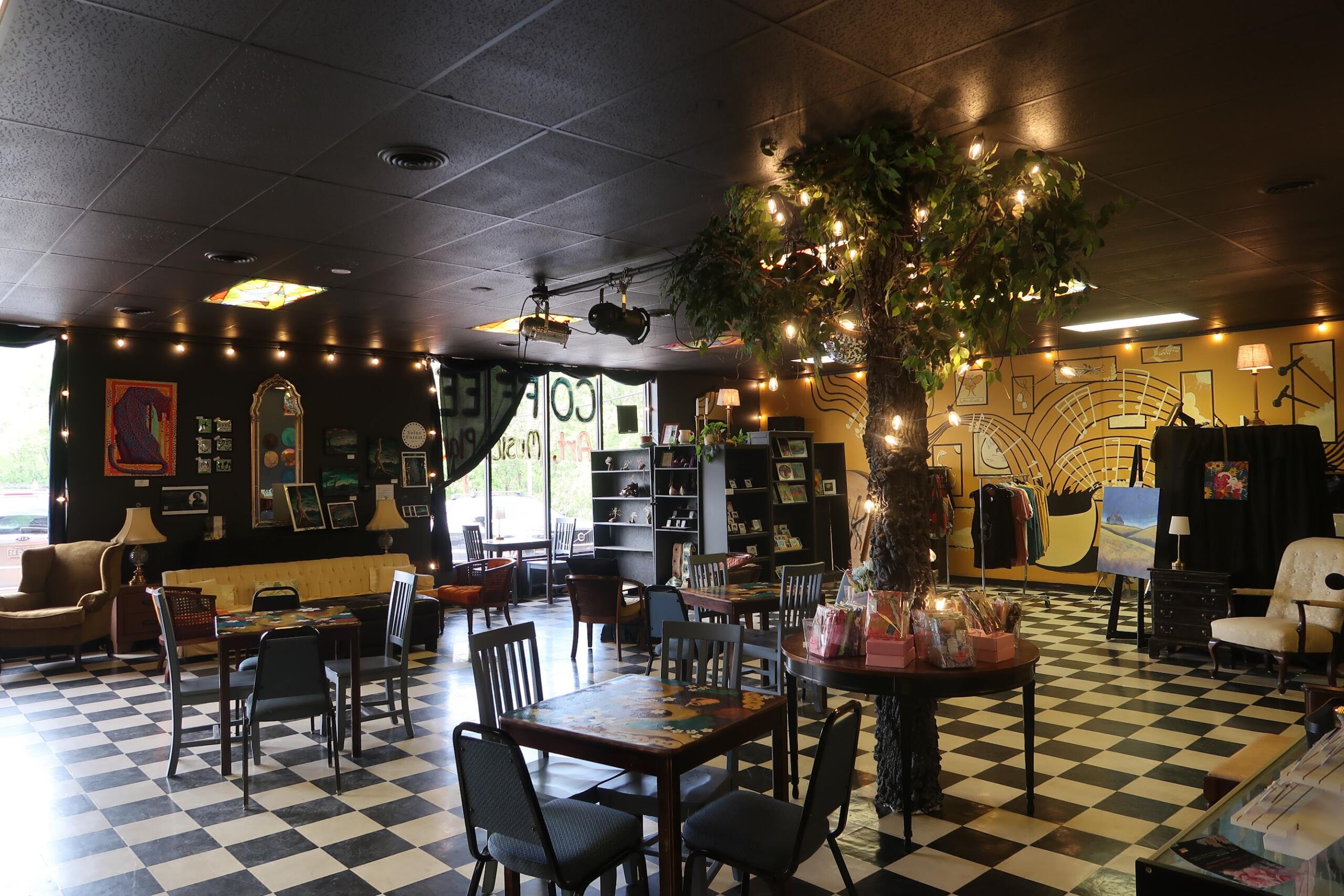 Interior of a cozy cafe/gallery with a black-and-white checker floor, string lights, and a large illuminated tree centerpiece.