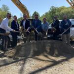 Group of professionals in suits and business attire shovel dirt at a construction site, with a large yellow excavator in the background.