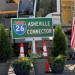Green I-26 Asheville Connector road sign with construction equipment in background and American flag nearby, in a construction site setting