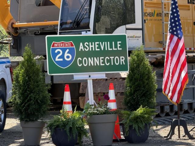 Green I-26 Asheville Connector road sign with construction equipment in background and American flag nearby, in a construction site setting