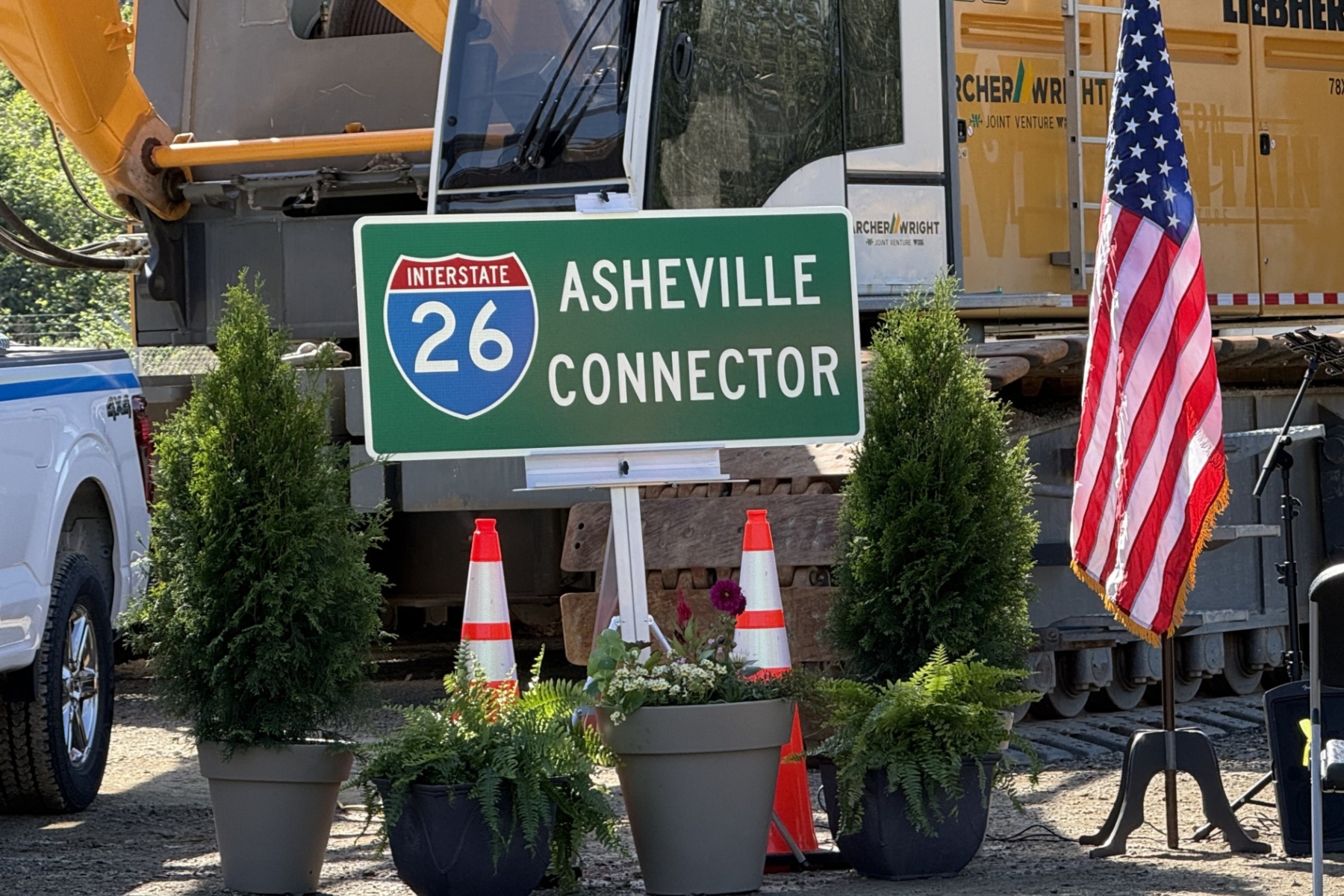 Green I-26 Asheville Connector road sign with construction equipment in background and American flag nearby, in a construction site setting