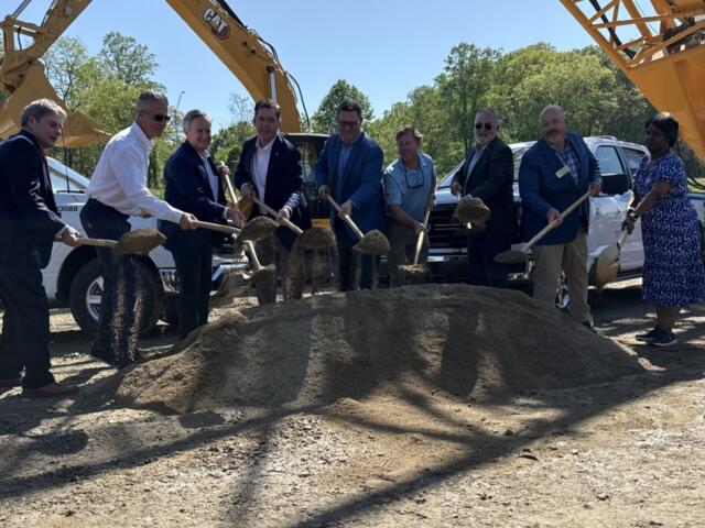Group of professionals in suits and business attire shovel dirt at a construction site, with a large yellow excavator in the background.