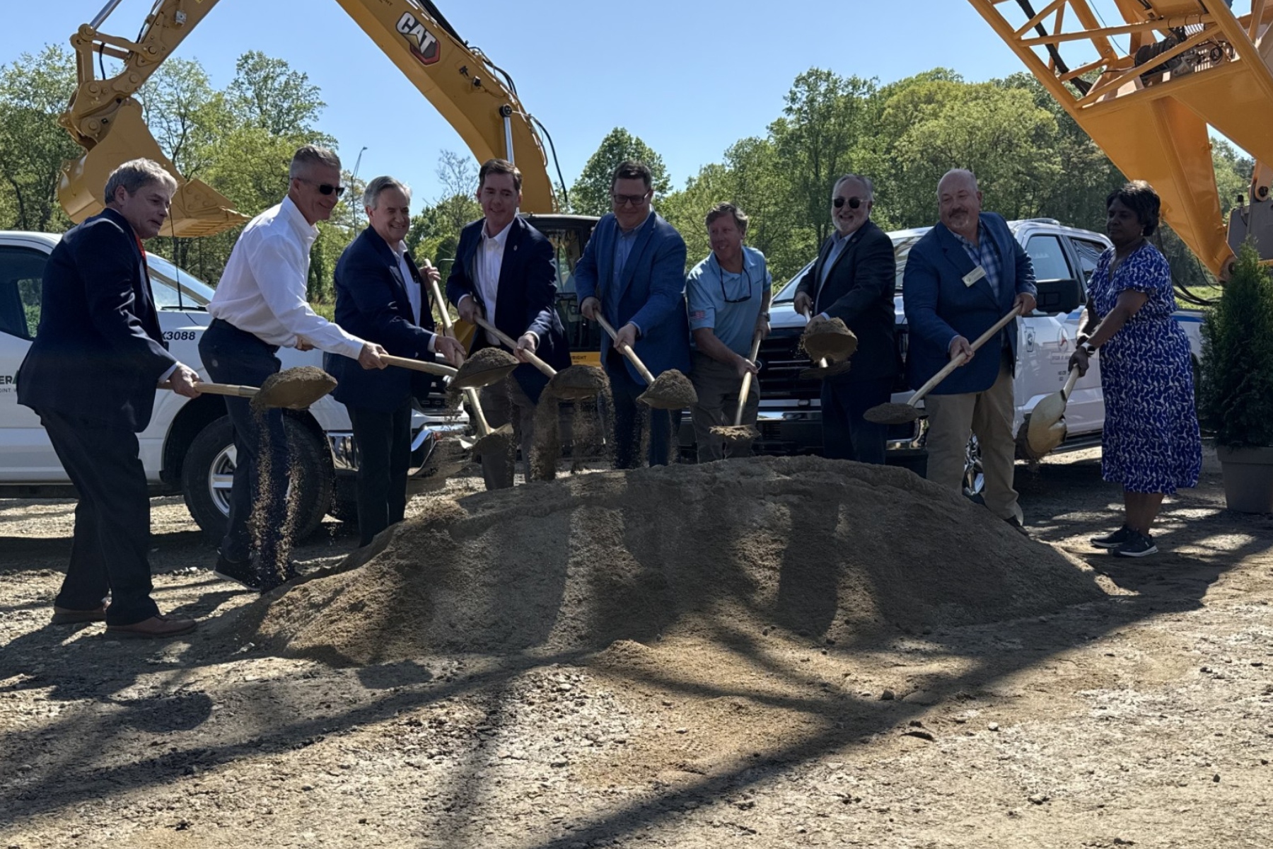 Group of professionals in suits and business attire shovel dirt at a construction site, with a large yellow excavator in the background.