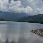 Calm lake with forested mountains in the distance and a rocky jetty on the right under a cloudy sky.