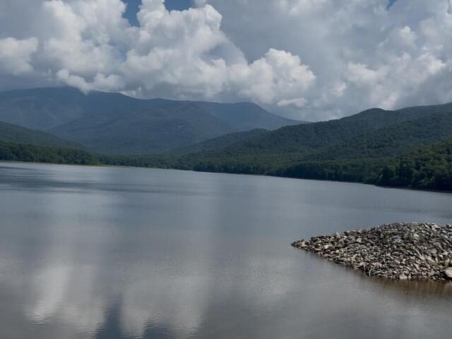Calm lake with forested mountains in the distance and a rocky jetty on the right under a cloudy sky.