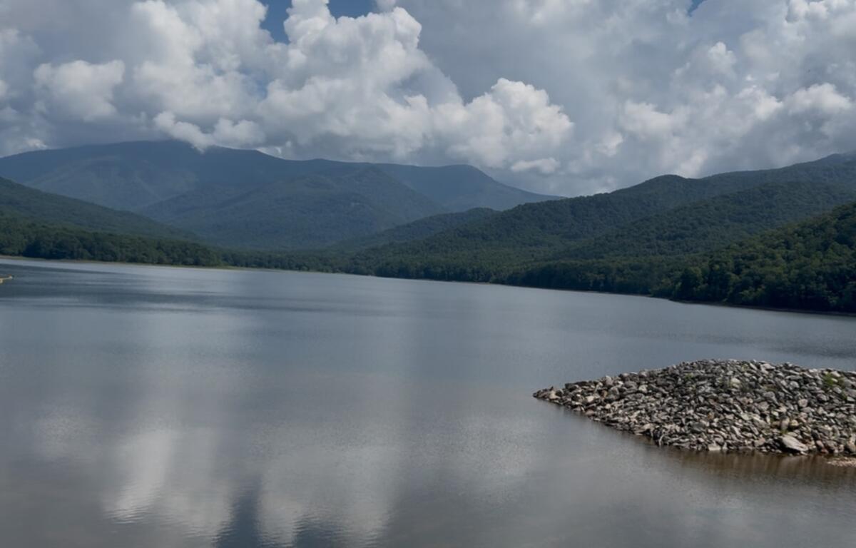 Calm lake with forested mountains in the distance and a rocky jetty on the right under a cloudy sky.