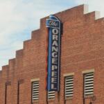 Brick building with a tall blue neon sign mounted along the corner, the sign featuring the word 'The' at the top and extending downward along the wall.