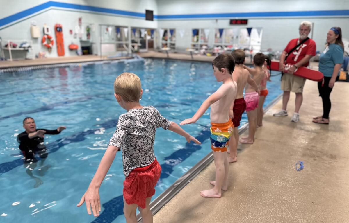 Children jump into a pool from the edge as a lifeguard and another adult supervise nearby indoors.