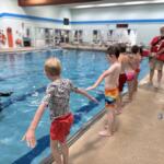 Children jump into a pool from the edge as a lifeguard and another adult supervise nearby indoors.