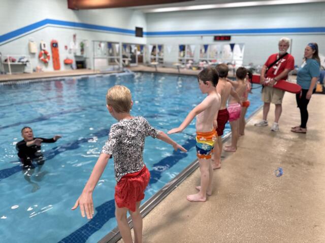 Children jump into a pool from the edge as a lifeguard and another adult supervise nearby indoors.
