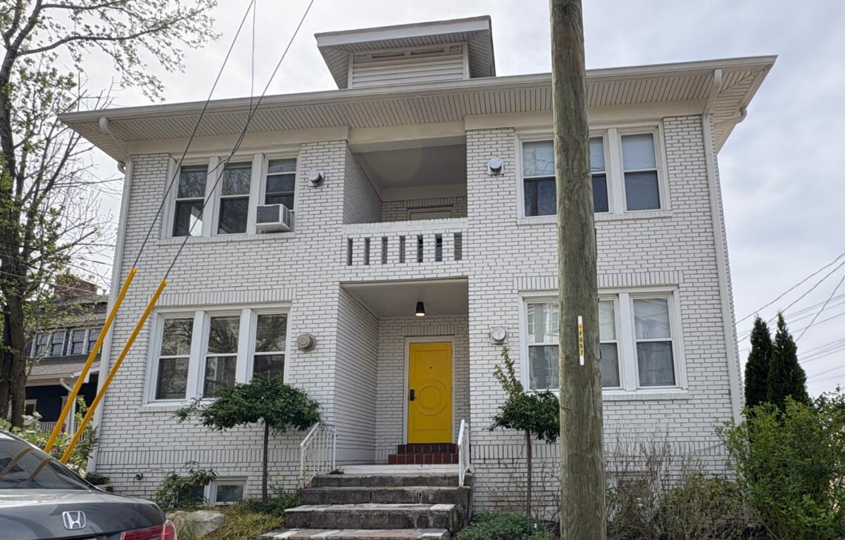 White brick apartment building with a bright yellow front door and brick steps leading up to it, greenery on both sides, and a utility pole in the foreground.