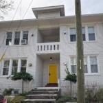 White brick apartment building with a bright yellow front door and brick steps leading up to it, greenery on both sides, and a utility pole in the foreground.
