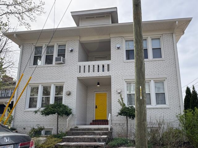 White brick apartment building with a bright yellow front door and brick steps leading up to it, greenery on both sides, and a utility pole in the foreground.