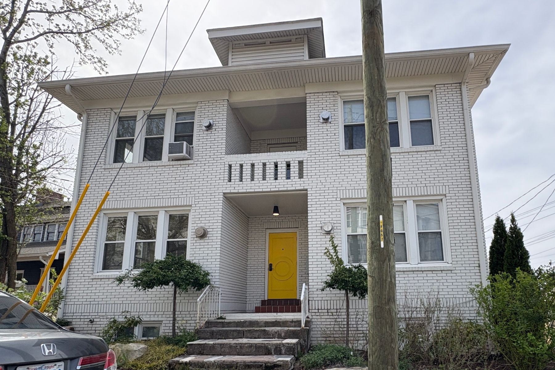White brick apartment building with a bright yellow front door and brick steps leading up to it, greenery on both sides, and a utility pole in the foreground.