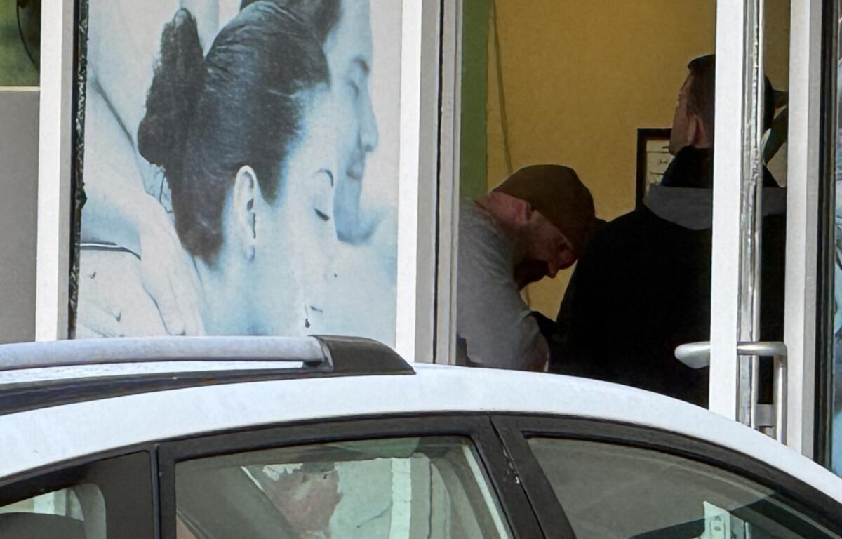 Storefront window with a large blue portrait of a woman on the left, a car in front, and two people inside the yellow-lit space on the right (one bowing, one wearing a headscarf).