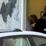 Storefront window with a large blue portrait of a woman on the left, a car in front, and two people inside the yellow-lit space on the right (one bowing, one wearing a headscarf).