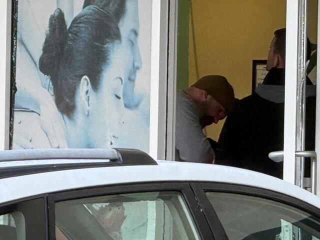Storefront window with a large blue portrait of a woman on the left, a car in front, and two people inside the yellow-lit space on the right (one bowing, one wearing a headscarf).