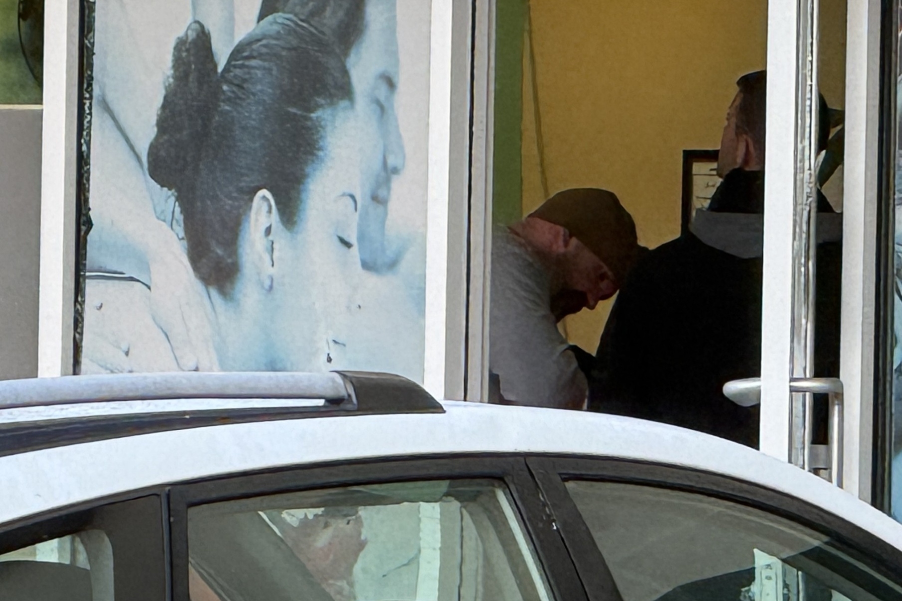 Storefront window with a large blue portrait of a woman on the left, a car in front, and two people inside the yellow-lit space on the right (one bowing, one wearing a headscarf).