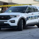 White Ford police SUV parked in front of a building with yellow police tape across the scene, 'POLICE' graphic on the side.