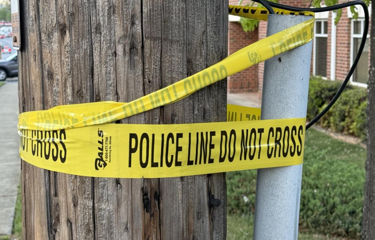 Yellow police line tape reading 'DO NOT CROSS' wrapped around a wooden utility pole and a nearby metal post, cordoning off a sidewalk area in a residential street.