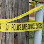 Yellow police line tape reading 'DO NOT CROSS' wrapped around a wooden utility pole and a nearby metal post, cordoning off a sidewalk area in a residential street.