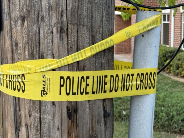 Yellow police line tape reading 'DO NOT CROSS' wrapped around a wooden utility pole and a nearby metal post, cordoning off a sidewalk area in a residential street.
