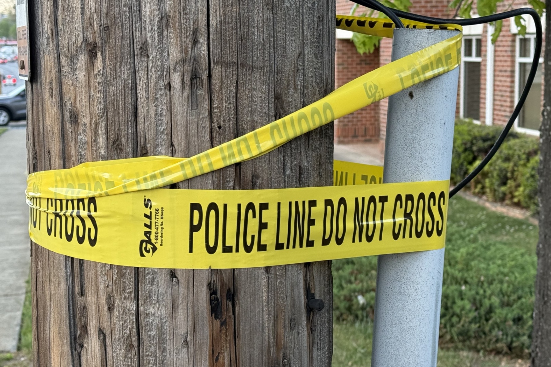 Yellow police line tape reading 'DO NOT CROSS' wrapped around a wooden utility pole and a nearby metal post, cordoning off a sidewalk area in a residential street.