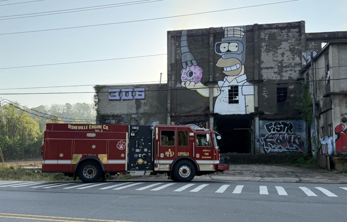 Red Asheville Fire Department fire engine parked in front of a graffiti-covered building with a large Simpsons-style mural holding a pink donut? donut is missing, large hand and Donut cartoon on wall